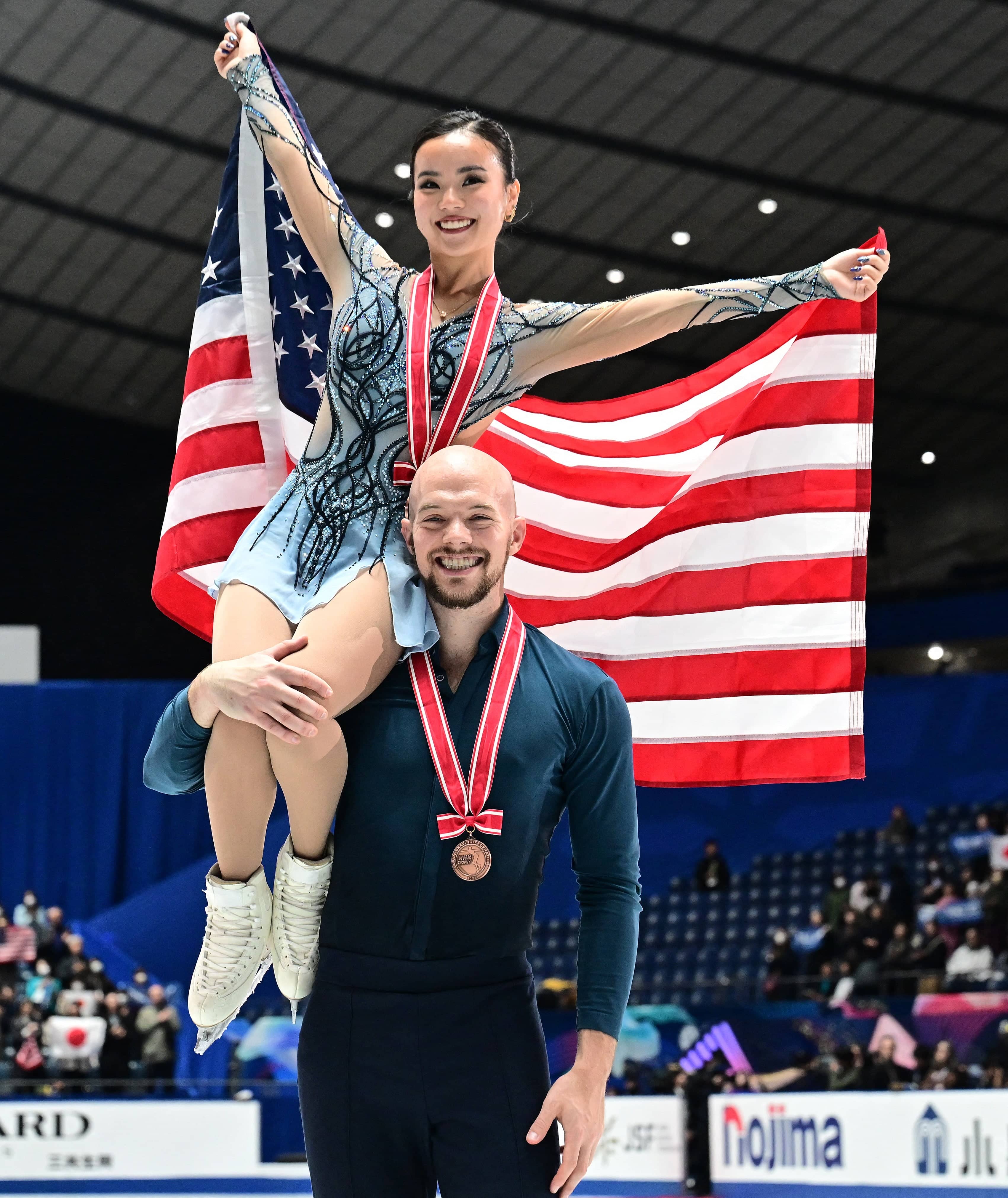 Ellie Kam sits on Danny O'Shea's shoulders, doing a lift during the victory ceremony of the pairs event at NHK Trophy. Both smile with bronze medals around their neck, and Ellie holds the U.S. flag behind her.